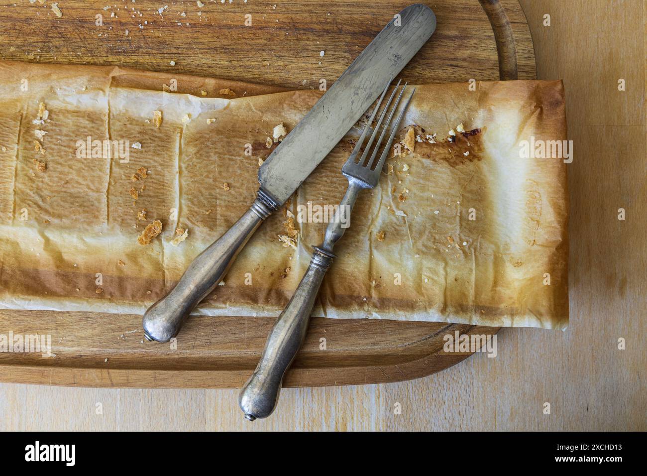 Photographie rapprochée sur table d'une planche à découper en bois d'acacia avec du papier sulfurisé laissé sur la tarte et fourchette vintage et couteau sur une table en bois Banque D'Images