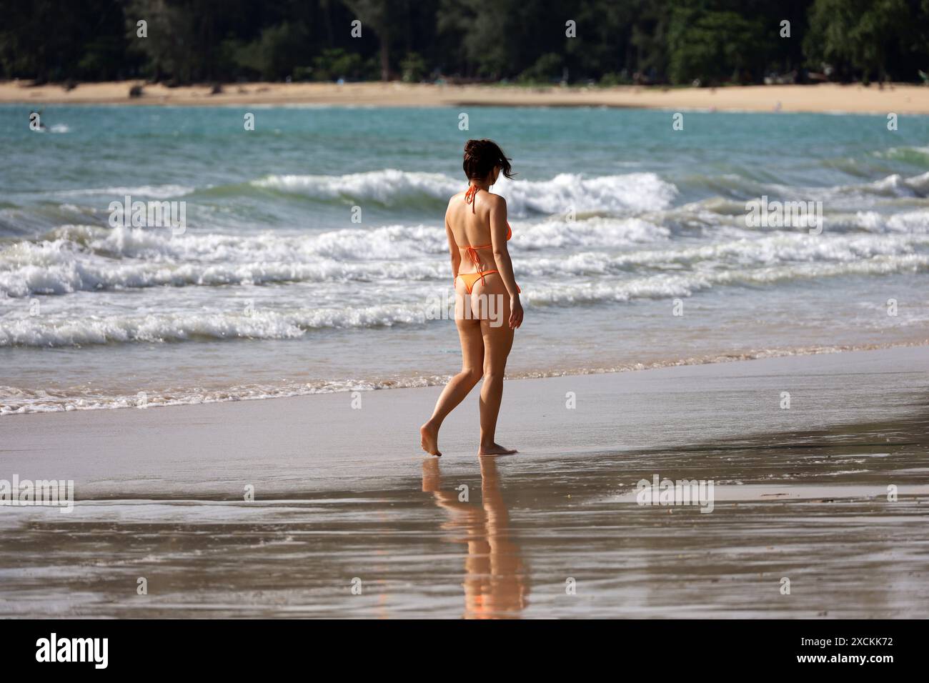 Femme en bikini orange marchant sur la plage de sable humide sur fond de vagues de mer. Profiter du soleil sur la côte tropicale Banque D'Images