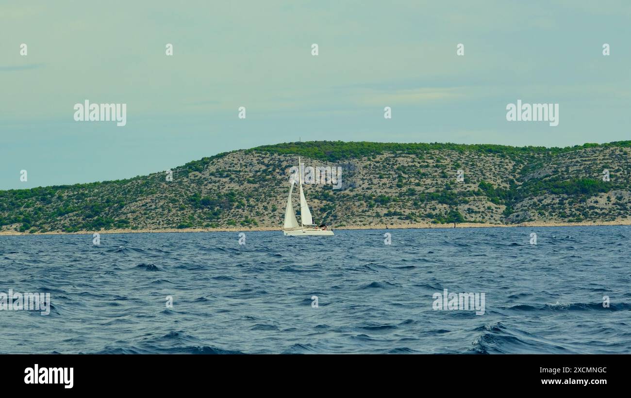 Un voilier navigue dans l'océan près d'une rive rocheuse. Le ciel est nuageux et l'eau est calme Banque D'Images