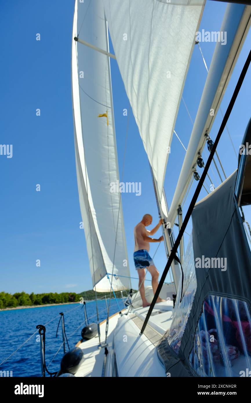 Un homme est debout sur un voilier, ajustant la voile. Le ciel est clair et bleu, et l'eau est calme. Concept de détente et de loisirs, comme l'homme e Banque D'Images