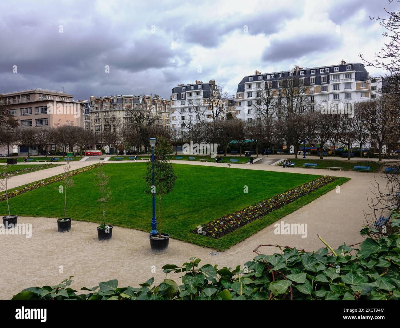 Des bâtiments de style parisien entourent la place Saint-Lambert tandis que les gens se détendent sur les bancs du parc par une journée nuageuse de mars dans le 15ème arrondissement, Paris, France. Banque D'Images