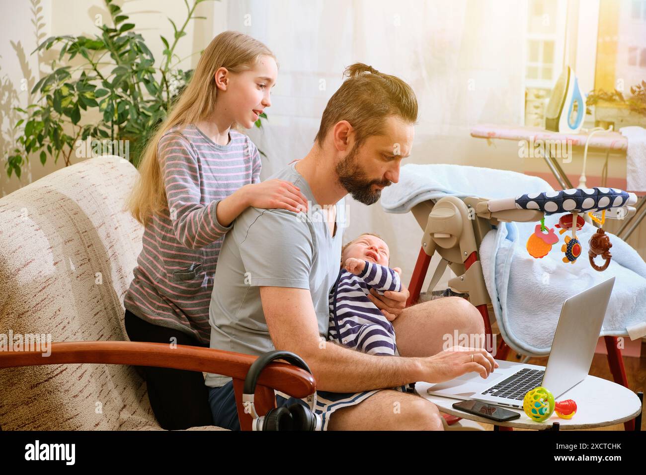 Père Caucasien multitâches comme il travaille sur un ordinateur portable, tient un bébé et est assisté par sa fille aînée dans un cadre confortable à la maison. Concept de bureau à domicile Banque D'Images