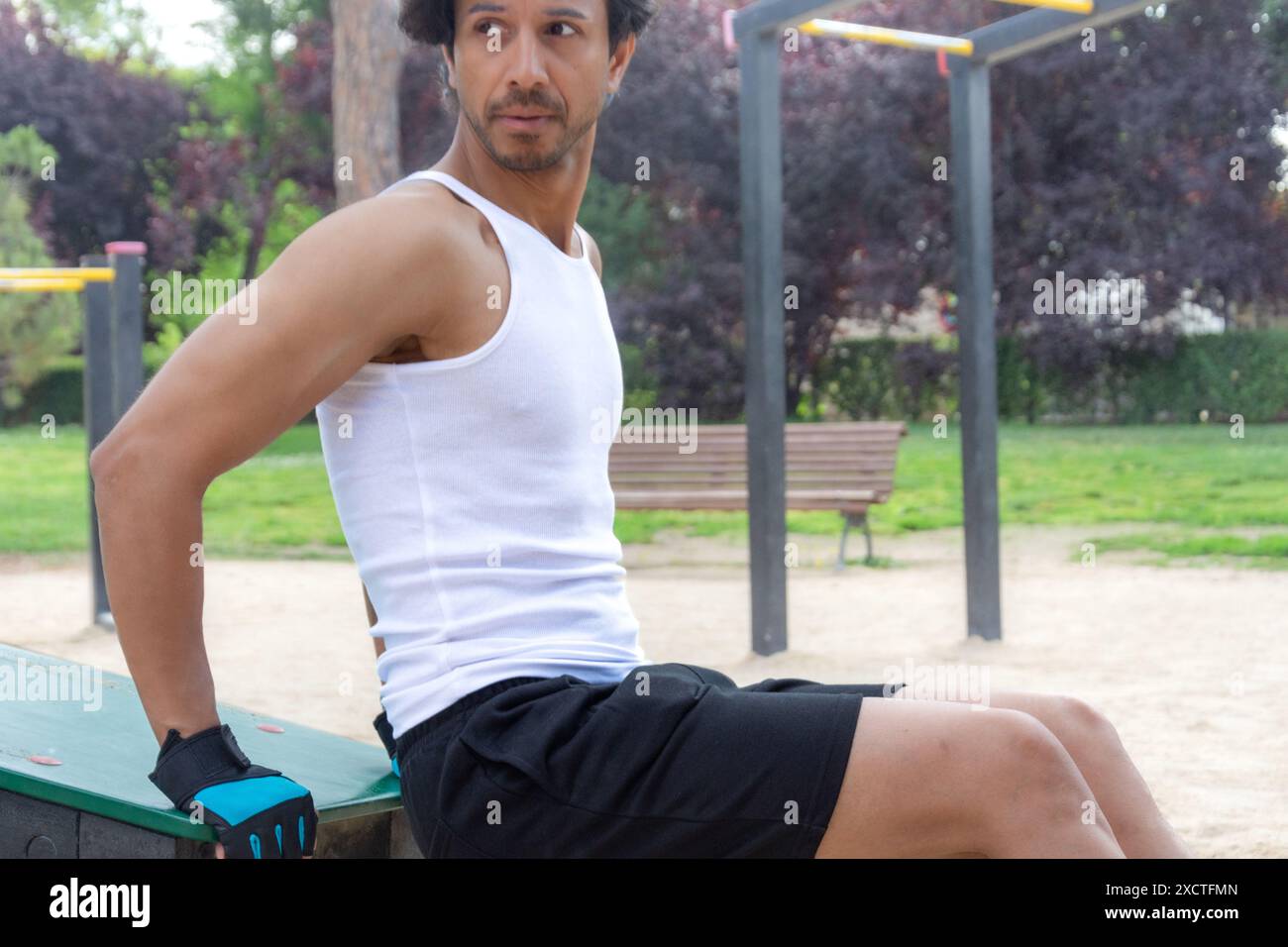 Un homme dans un débardeur blanc est assis sur un banc dans un parc. Le banc est à côté d'une structure métallique avec un poteau jaune Banque D'Images Un homme dans un débardeur blanc est assis sur un banc dans un parc. Le banc est à côté d'une structure métallique avec un poteau jaune Banque D'Images