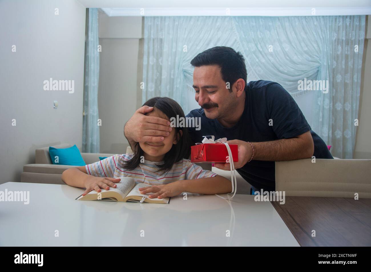 Le père qui ferme les yeux de sa fille pendant qu'elle lit un livre et lui donne un coffret cadeau rouge Banque D'Images