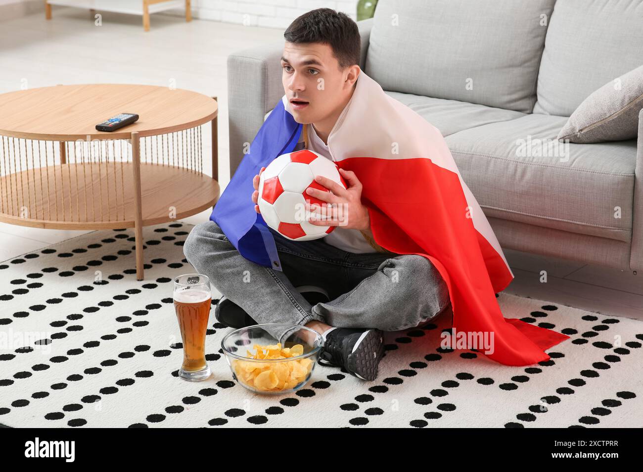 Jeune homme avec un verre de bière, ballon de football, bol de chips de pomme de terre et drapeau de la France regardant le football à la maison Banque D'Images