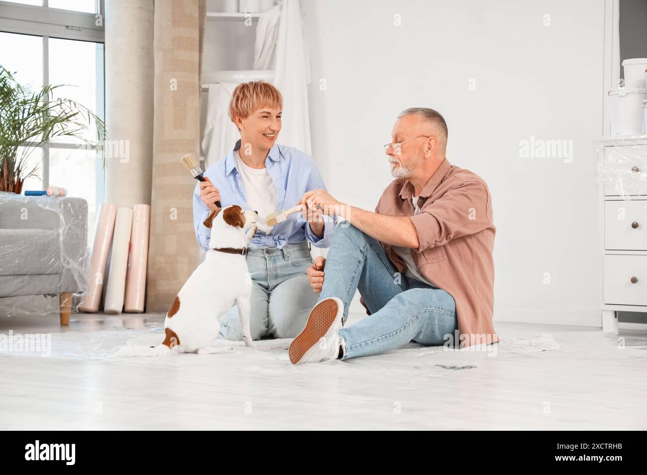 Couple mûr avec des pinceaux et chien assis sur le sol pendant la réparation dans leur nouvelle maison Banque D'Images