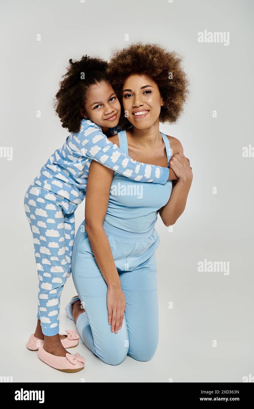 Embrassant amour et rire, une mère afro-américaine et sa fille prennent la pose dans un pyjama bleu assorti sur un fond gris. Banque D'Images