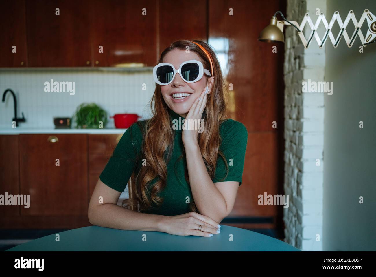 Femme rousse joyeuse dans une cuisine vintage de style années 50 posant avec de grandes lunettes de soleil blanches, regardant directement la caméra, ajoutant une touche humoristique à t Banque D'Images