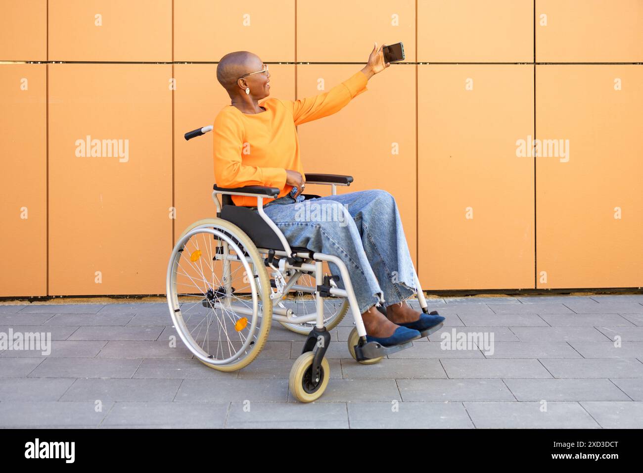 Une femme d'affaires afro-américaine en fauteuil roulant capture un selfie sur un fond orange moderne, mettant en valeur l'indépendance et l'étreinte technologique Banque D'Images