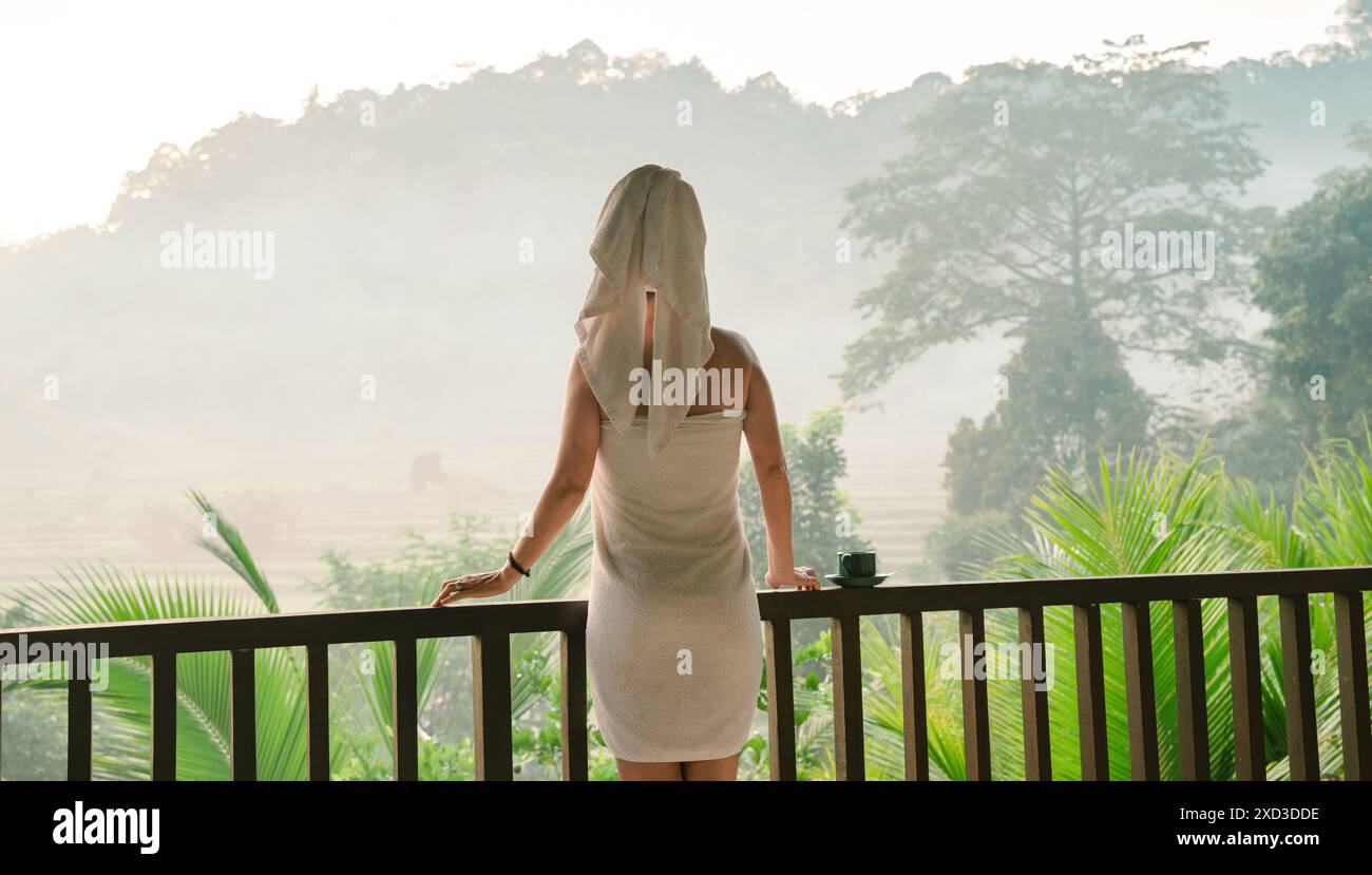Une femme dans une serviette se tient sur un balcon en bois, regardant la vue brumeuse et remplie d'arbres, avec une tasse de thé à côté d'elle, incarnant la paix et la tranquillité Banque D'Images