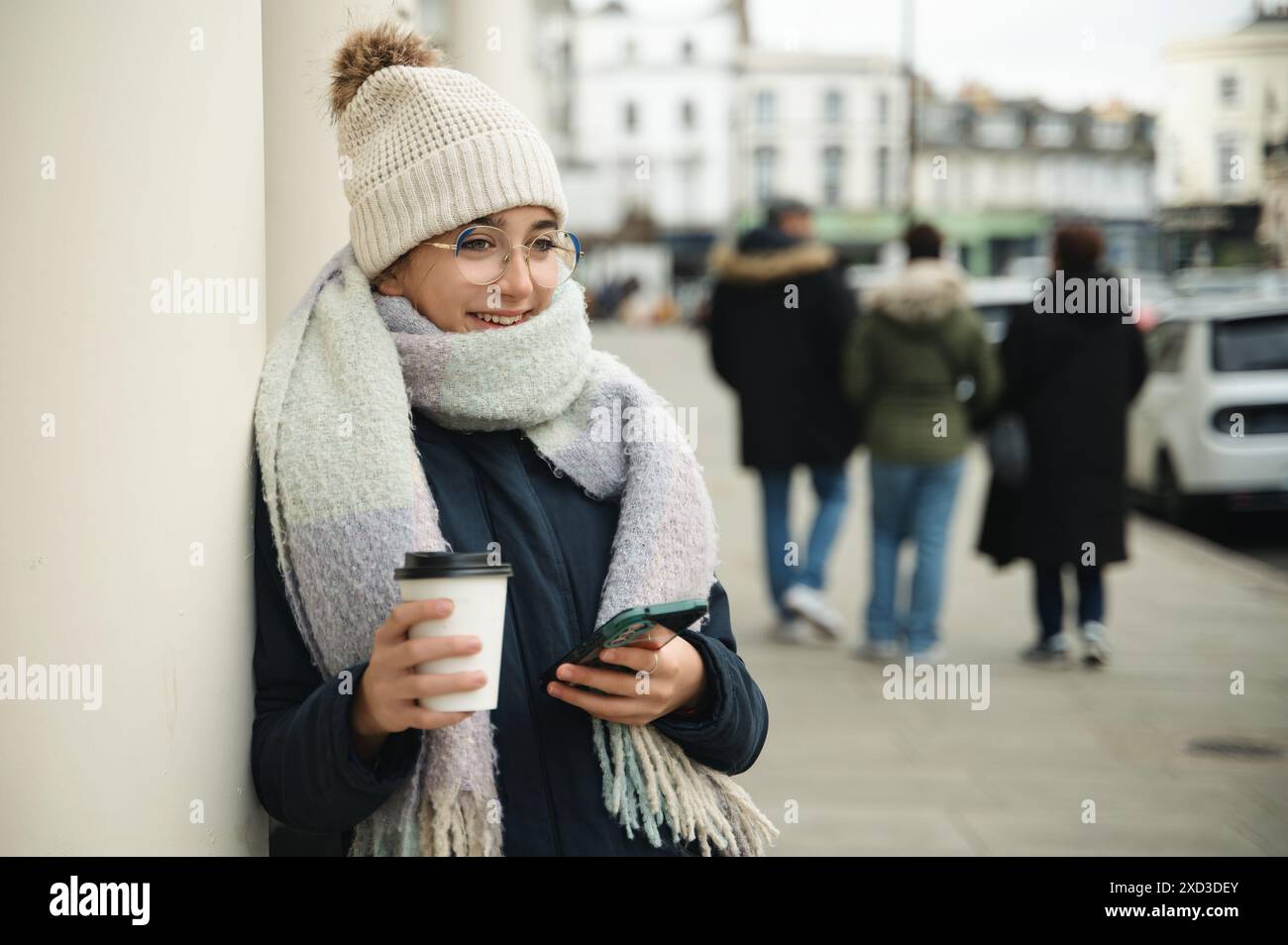 Jeune femme habillée à la mode profitant de son café tout en regardant son smartphone dans une rue animée de Londres. Banque D'Images