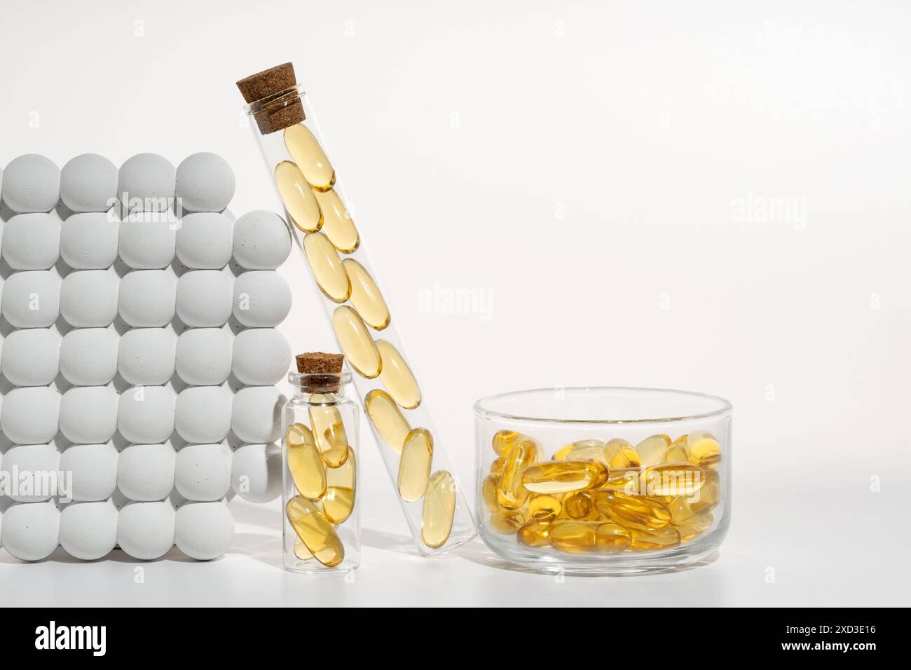 Un arrangement artistique de capsules d'huile de poisson oméga 3 présentées dans divers récipients en verre, y compris un flacon verseur et un bol, serrés contre un b blanc Banque D'Images