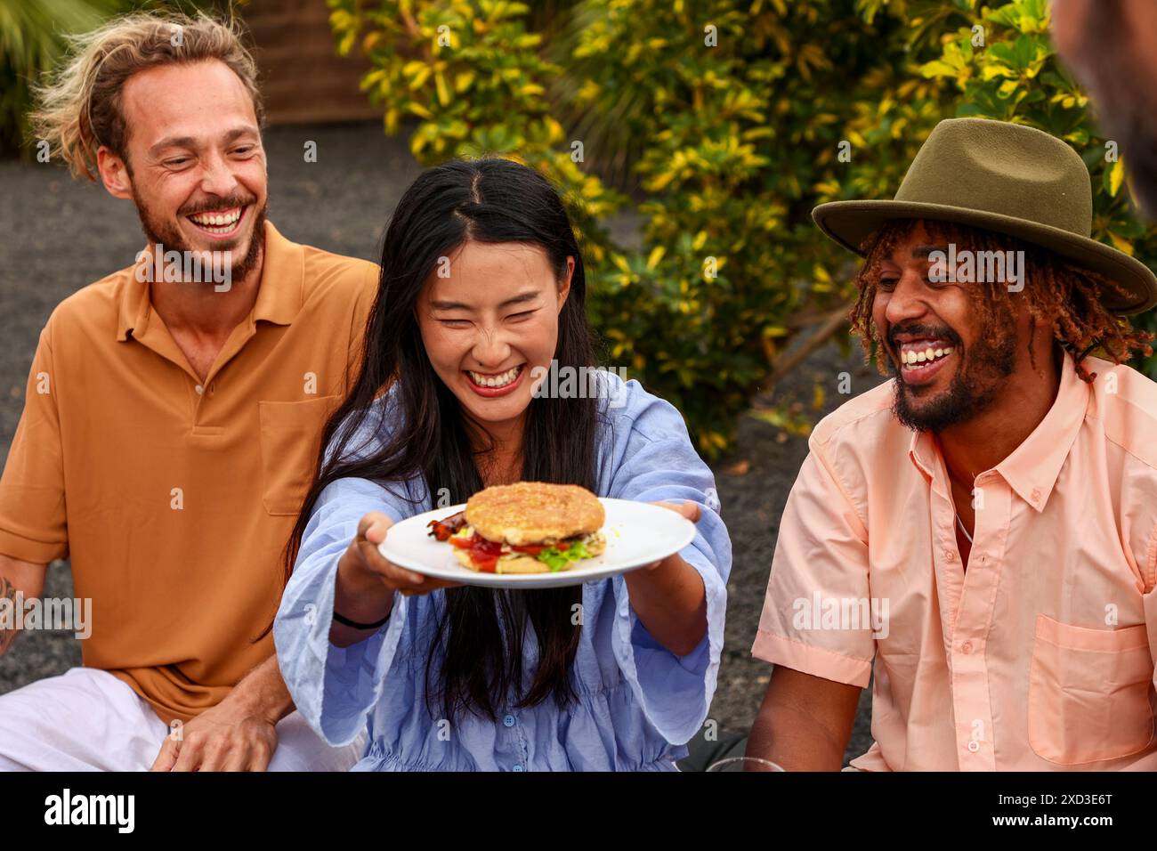 Trois amis joyeux partagent un rire à un barbecue dans la cour arrière. Une femme asiatique offre un hamburger à un homme caucasien et à un homme afro-américain. Tous sont casu Banque D'Images
