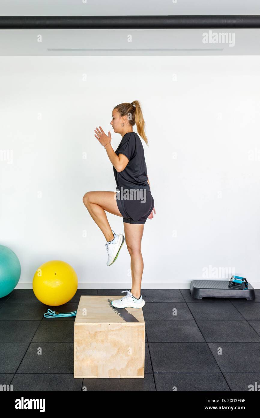 Une jeune femme en tenue sportive effectue un exercice de saut de boîte dynamique dans un gymnase contemporain et bien équipé, en se concentrant sur le renforcement de la force et de l'agili Banque D'Images