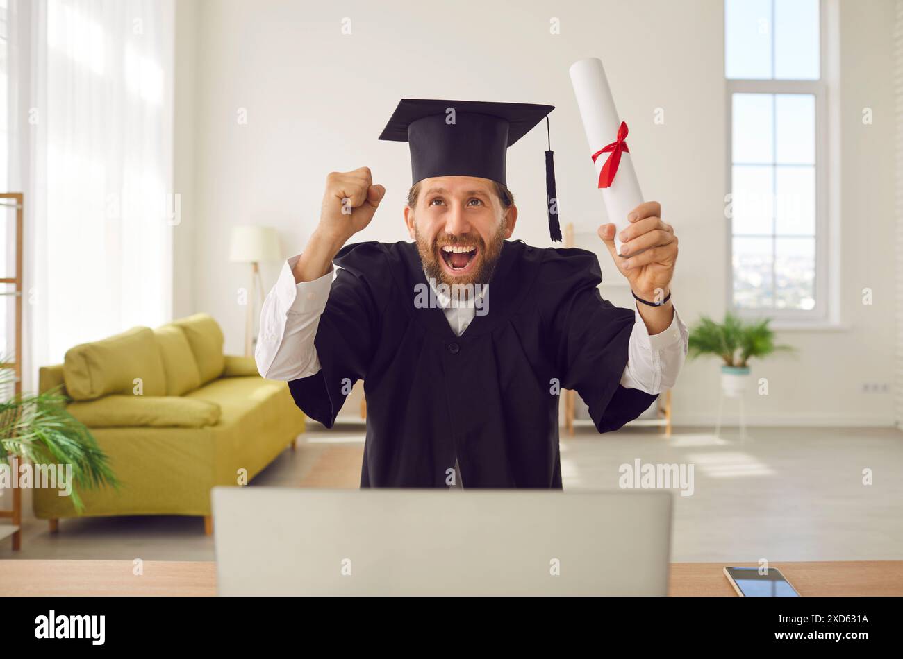 Homme dans la robe de gradué assis à la table avec diplôme faisant le geste oui pendant l'appel vidéo en ligne. Banque D'Images