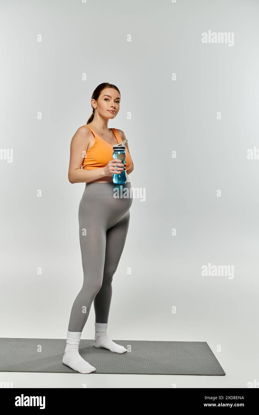 Femme enceinte sportive en tenue active tenant une bouteille d'eau tout en se tenant debout sur un tapis sur un fond gris. Banque D'Images