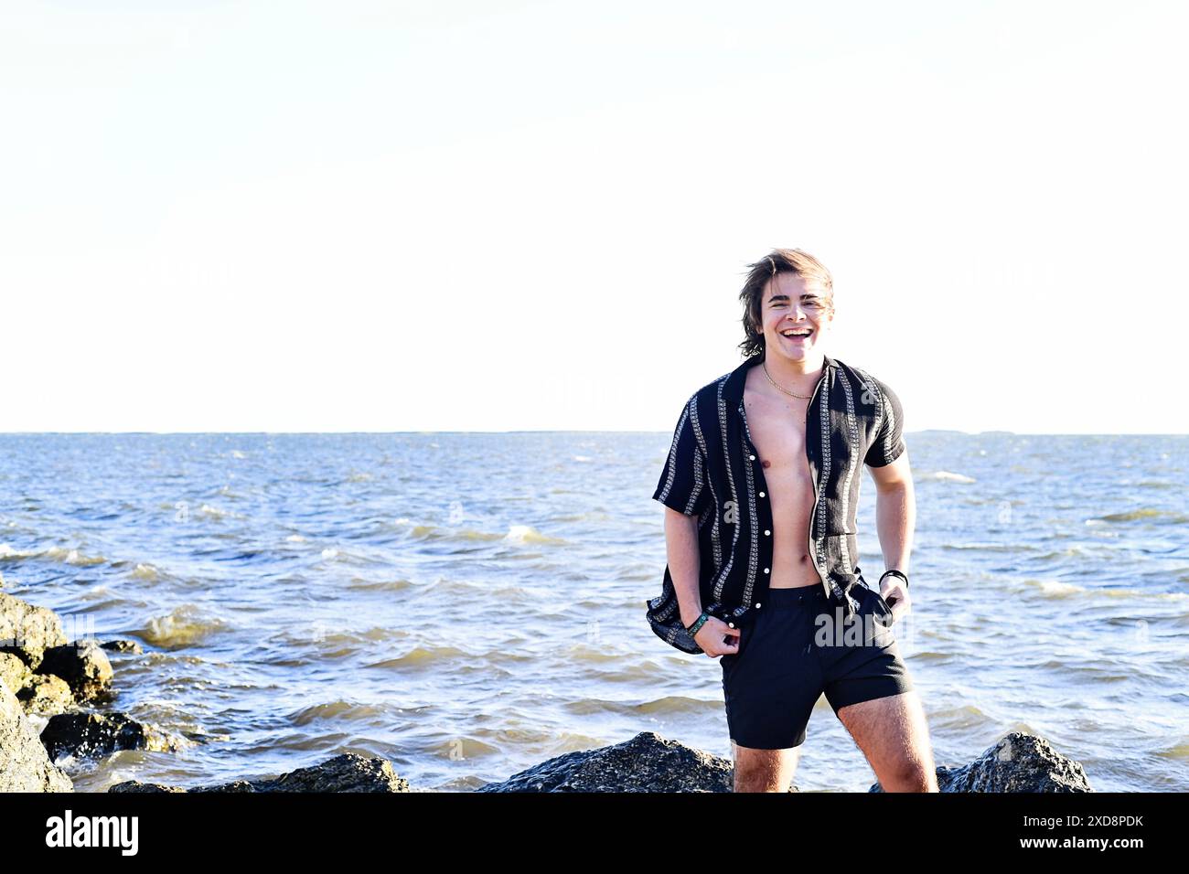 Jeune homme souriant debout sur des rochers près de l'océan, portant une op Banque D'Images