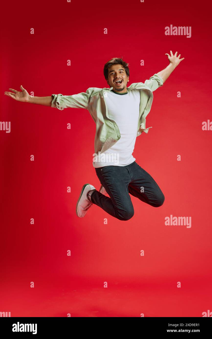 Photo dynamique de jeune homme indien sautant dans les airs de joie et de bonheur sur fond rouge de studio. Émotions positives. Banque D'Images