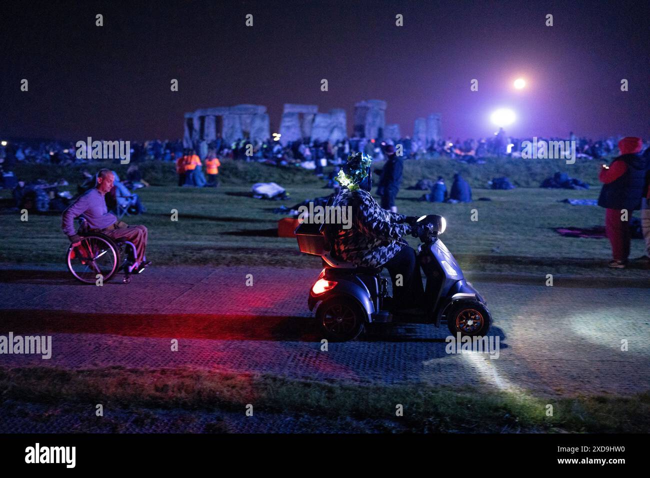 Un homme en fauteuil roulant et une femme utilisent un scooter de mobilité avec des fêtards avant l'aube célèbrent le solstice d'été (mi-été et jour le plus long) sur les anciennes pierres du néolithique tardif de Stonehenge, le 21 juin 2024, dans le Wiltshire, en Angleterre. Le solstice d'été est le jour le plus long de l'hémisphère nord et la nuit la plus courte de l'année, lorsque l'axe de la terre est incliné à son point le plus proche du soleil et les païens disent que l'ancien monument est un lieu sacré qui relie la Terre, la Lune, le Soleil et les saisons. Stonehenge a été construit en trois phases entre 3 000 av. J.-C. et 1 600 av. J.-C. Stonehenge est la propriété de H anglais Banque D'Images