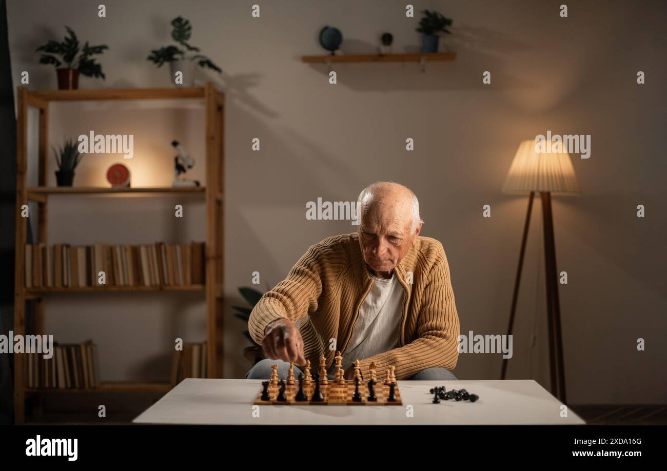 Vieil homme jouant aux échecs seul à table le soir à la maison. Banque D'Images