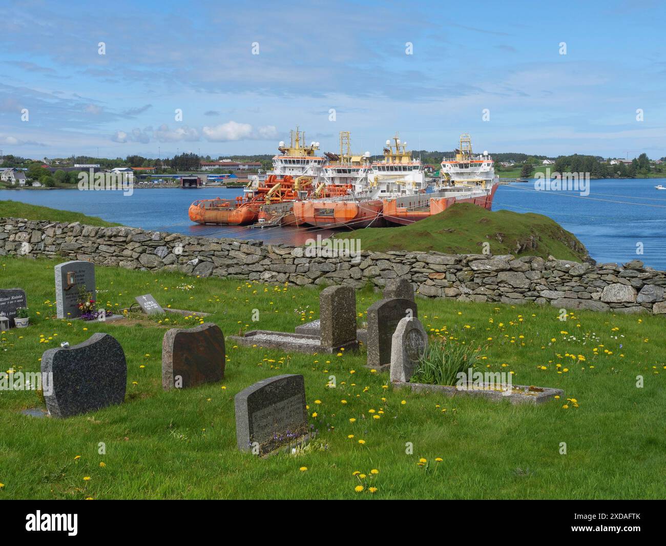 Vue d'un cimetière avec des pierres tombales et un mur de pierre au premier plan, derrière la mer avec plusieurs navires et un ciel bleu clair, haugesund, norvège Banque D'Images