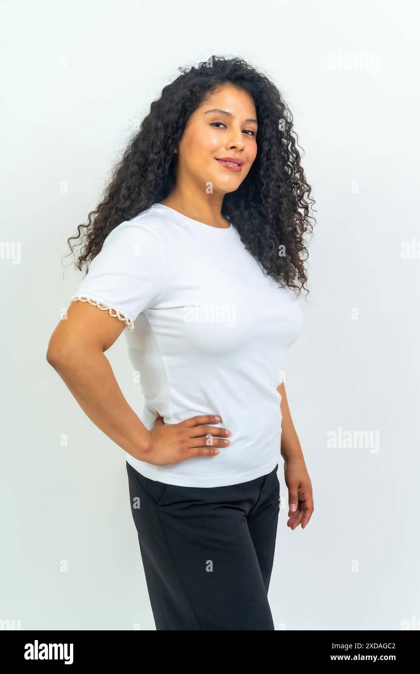 Portrait de studio avec fond blanc d'une femme latine avec des cheveux bouclés debout main fière sur la hanche souriant à la caméra Banque D'Images