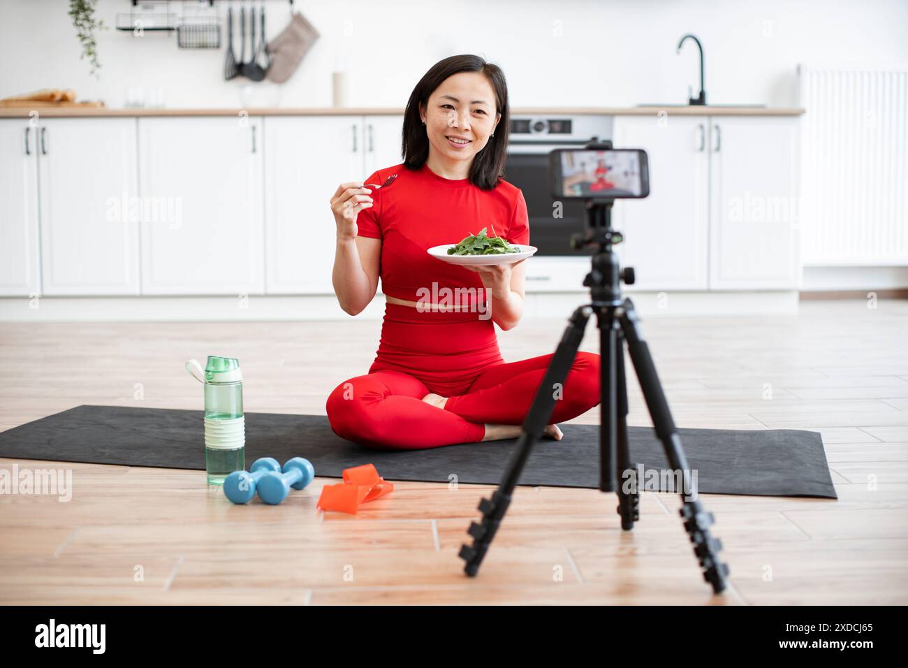 Femme en vêtements d'activité mangeant de la salade tout en enregistrant la vidéo de fitness dans la cuisine Banque D'Images