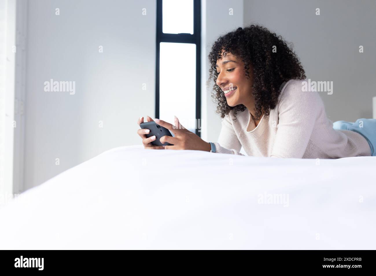 Femme allongée sur le lit tenant le smartphone, souriant et s'engageant avec l'appareil Banque D'Images