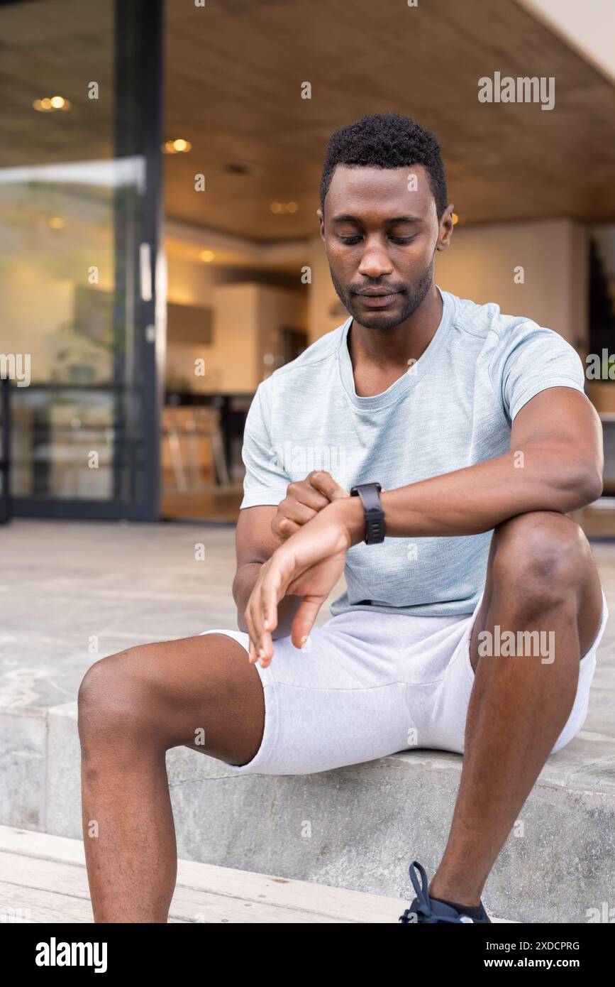 Homme afro-américain en vêtements décontractés vérifiant smartwatch tout en étant assis sur les marches Banque D'Images