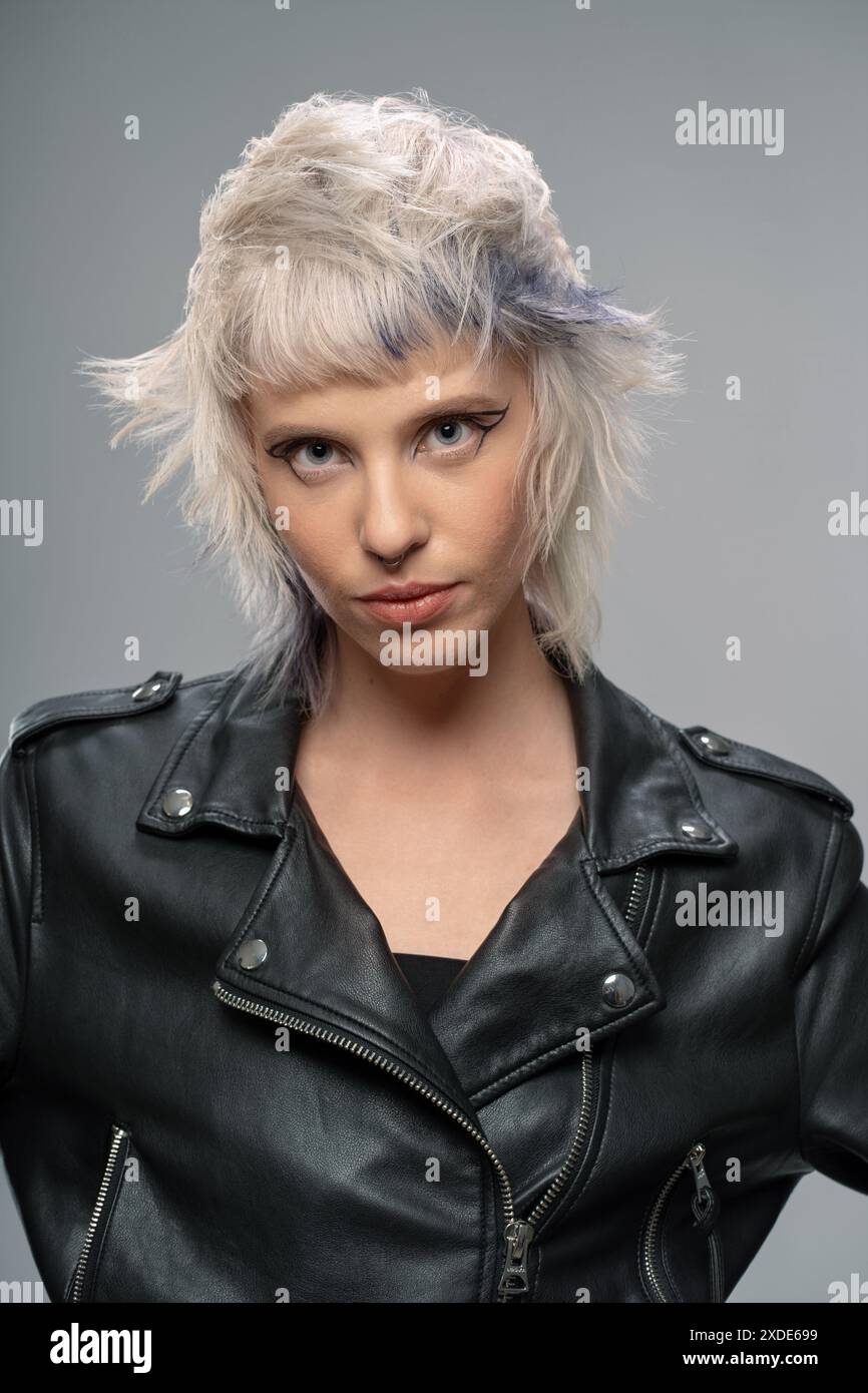 Une jeune femme avec une coiffure punk et une expression féroce, portant une veste en cuir, regardant l'appareil photo, fond gris, mode audacieuse, look moderne Banque D'Images