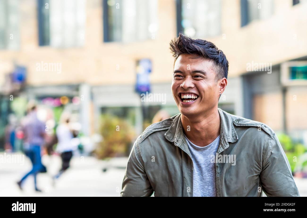 Jeune homme asiatique riant joyeusement dans un cadre urbain avec un fond de ville flou. L'image capture un moment de bonheur authentique et spontané Banque D'Images