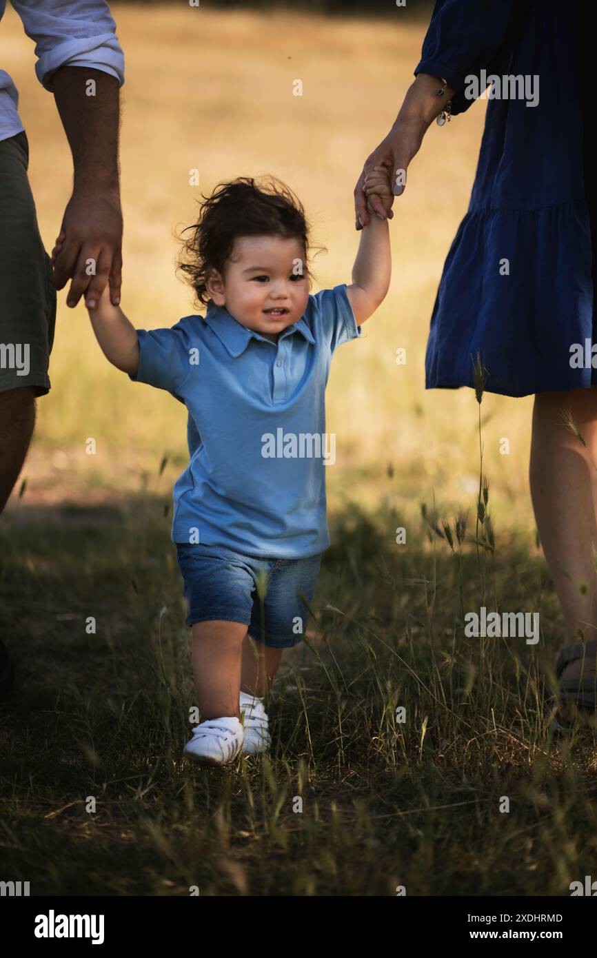 Un garçon d'un an marchant sur l'herbe, tenant les mains de ses parents, portant une chemise bleue et un short en Jean bleu. Banque D'Images