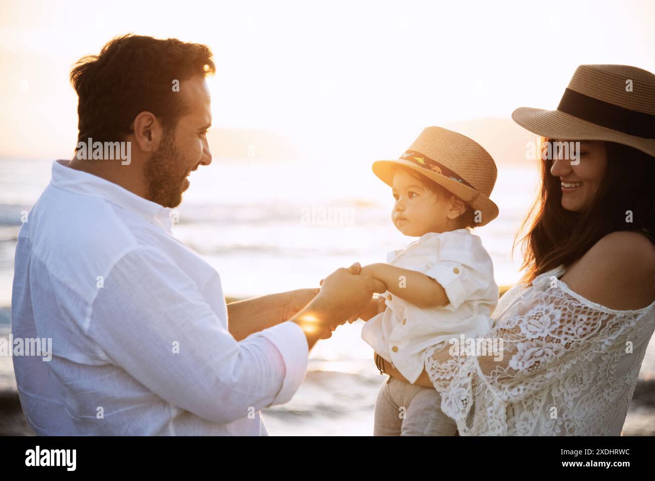 Un garçon d’un an portant une chemise blanche, des lunettes de soleil et un chapeau interagit avec ses parents sur la plage. La toile de fond comprend la mer et un beau coucher de soleil Banque D'Images