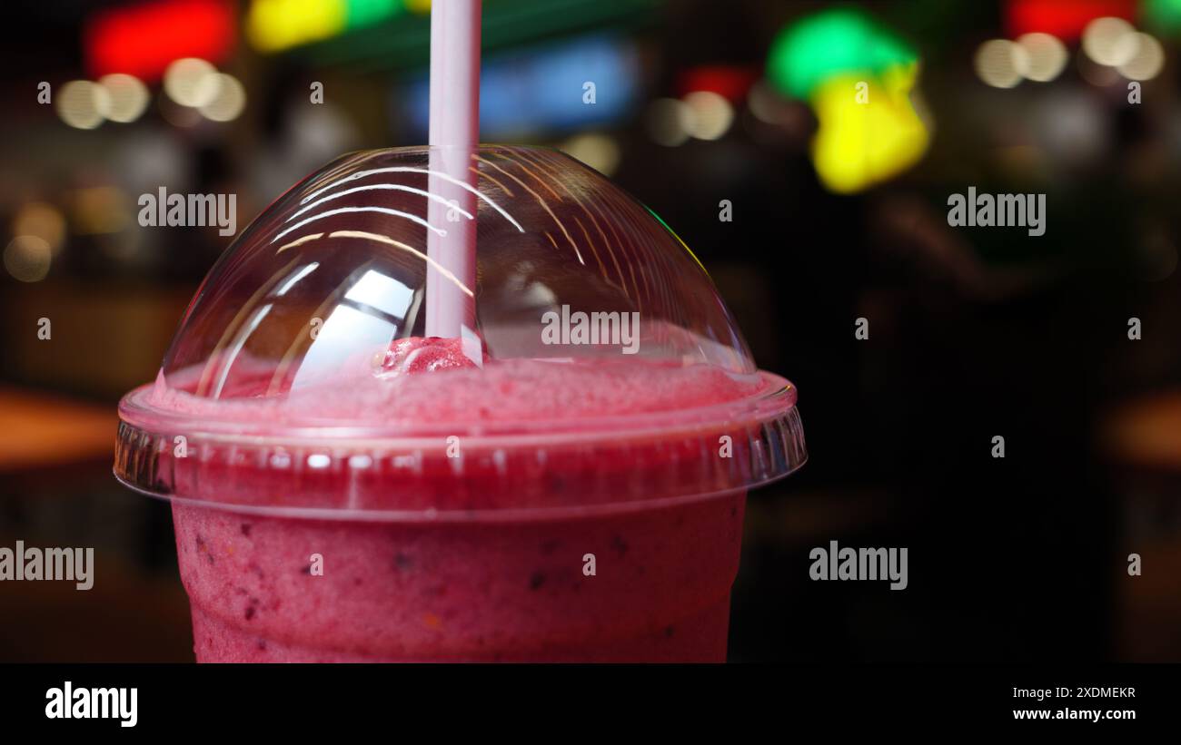 Un gros plan d'un smoothie rose dans une tasse en plastique avec une paille, dans un cadre de café lumineux. Le smoothie est recouvert d'un couvercle en dôme en plastique transparent. Banque D'Images