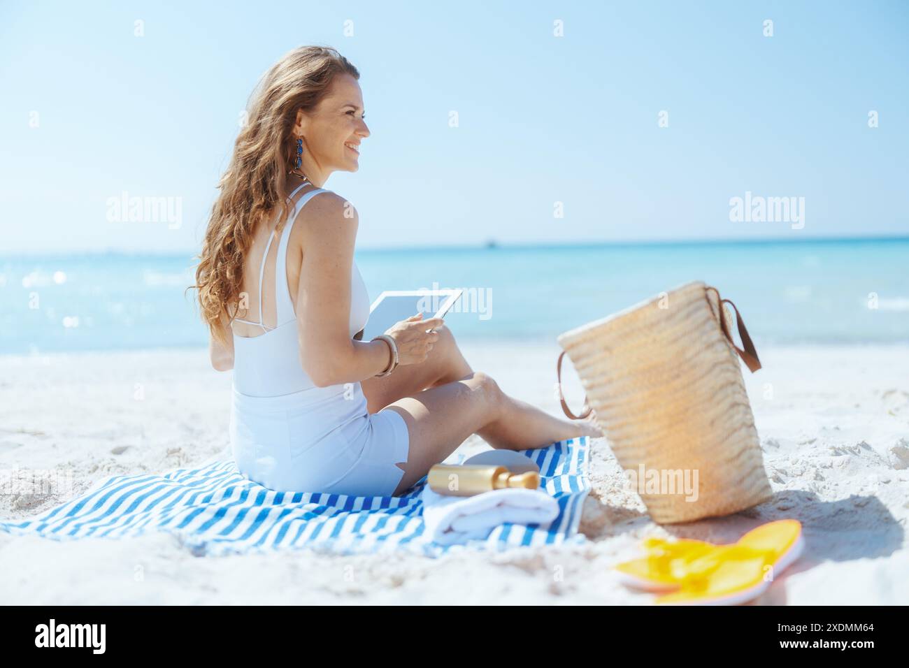 Femme élégante souriante sur la rive de l'océan avec sac de paille et serviette rayée en utilisant des applications sur tablette PC. Banque D'Images