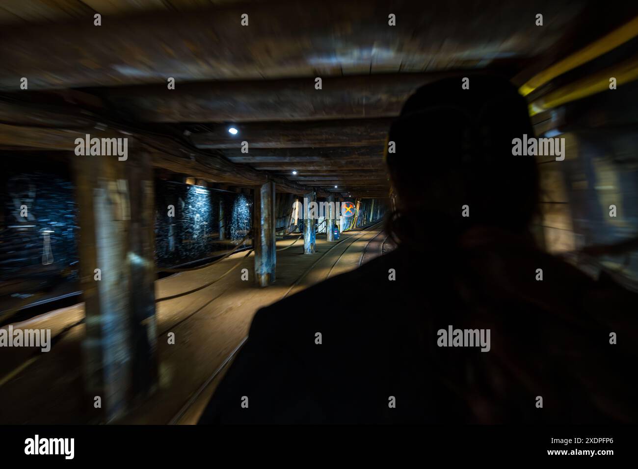 Des étais en bois soutiennent le plafond du tunnel aux Salzwelten Hallein tandis que le chemin de fer de la mine transporte les visiteurs profondément dans le Dürrnberg près de Hallein, Salzbourg, Autriche. Au premier plan à droite, la silhouette floue d'un passager est visible. Une lumière orange brille en arrière-plan. Banque D'Images