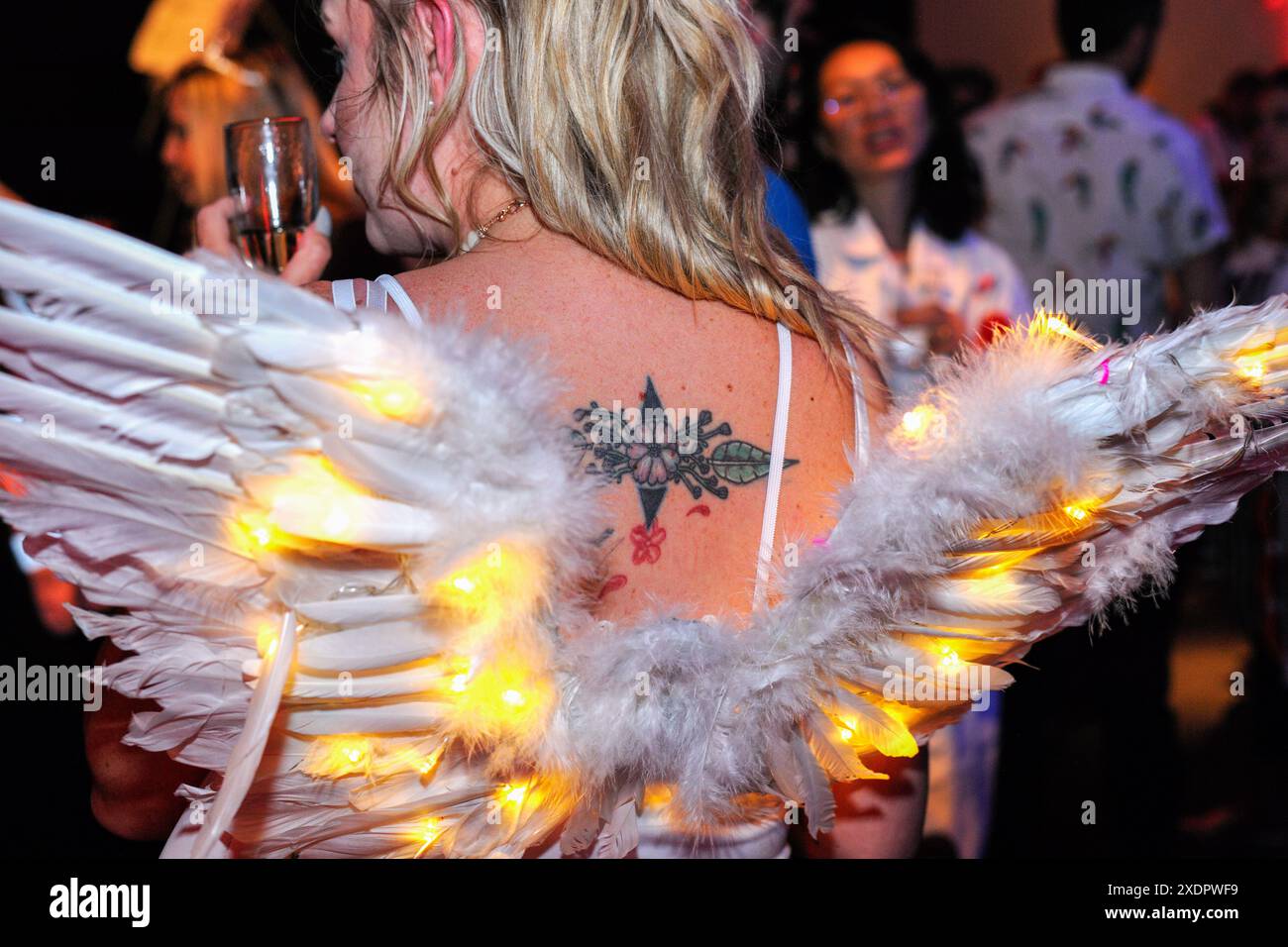 Jeune femme avec un tatouage sur le dos, portant des ailes d'ange avec des lumières de fée, et une coupe de champagne à la main, lors d'une fête Banque D'Images