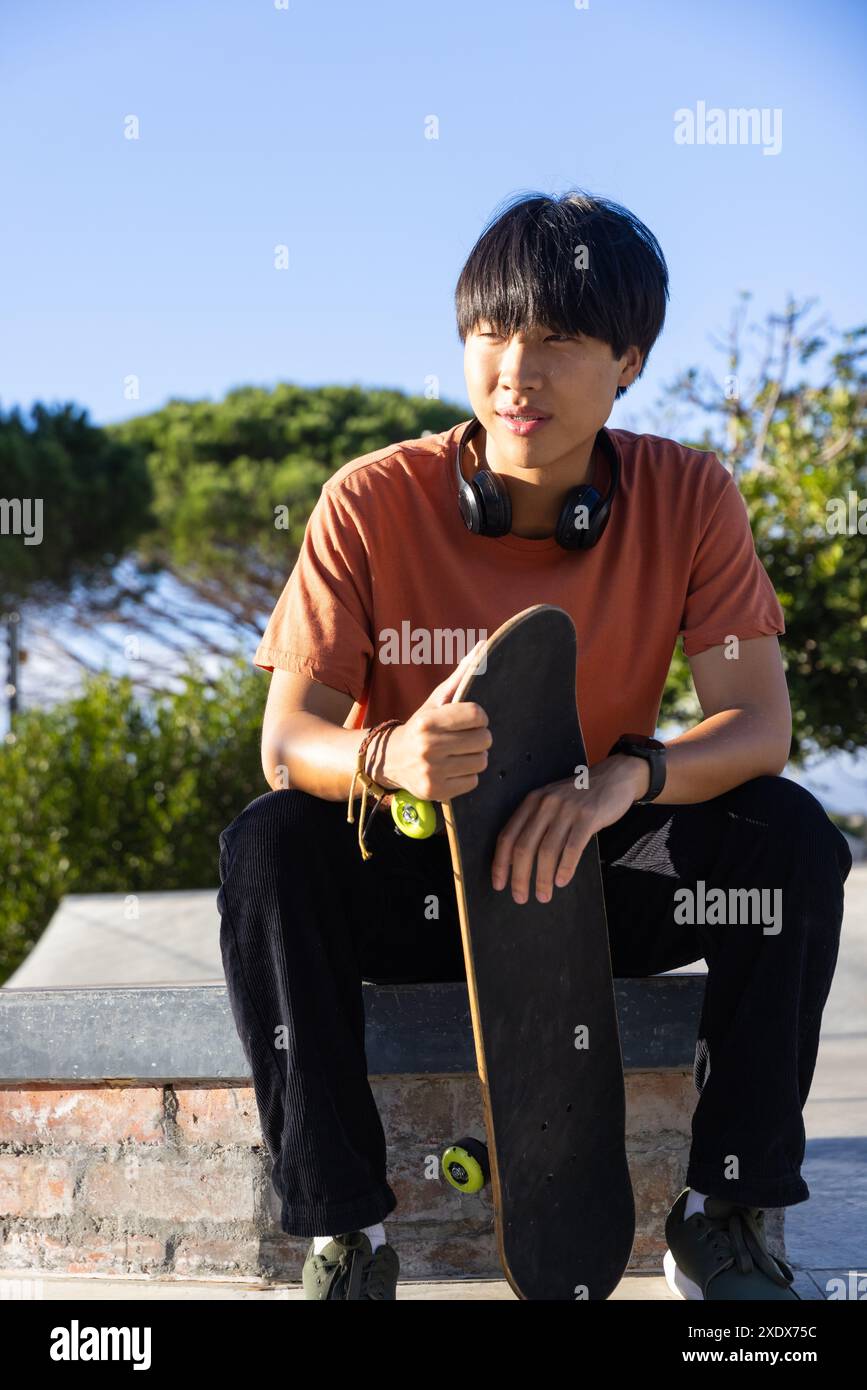 Tenant skateboard, adolescent assis à l'extérieur avec des écouteurs autour du cou Banque D'Images