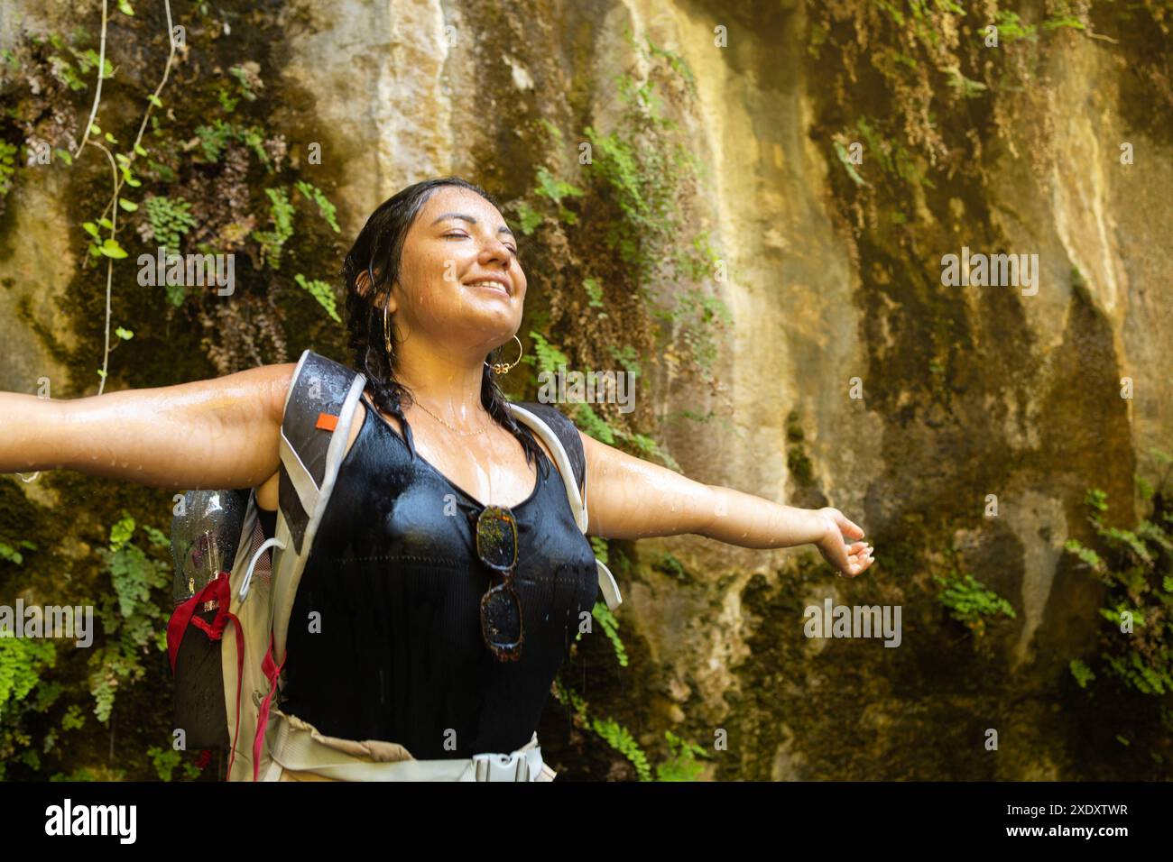 Une femme se tient debout les bras tendus sous une cascade rafraîchissante dans un canyon luxuriant. Elle porte un sac à dos et semble être sur une aventure de randonnée Banque D'Images