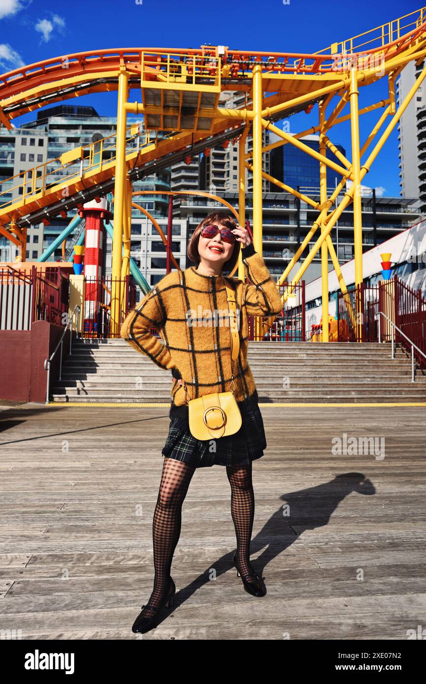 Une femme portant un pull à carreaux, une jupe et des bas à Luna Park sur le port de Sydney, une tenue jaune devant les montagnes russes rouges et jaunes Banque D'Images