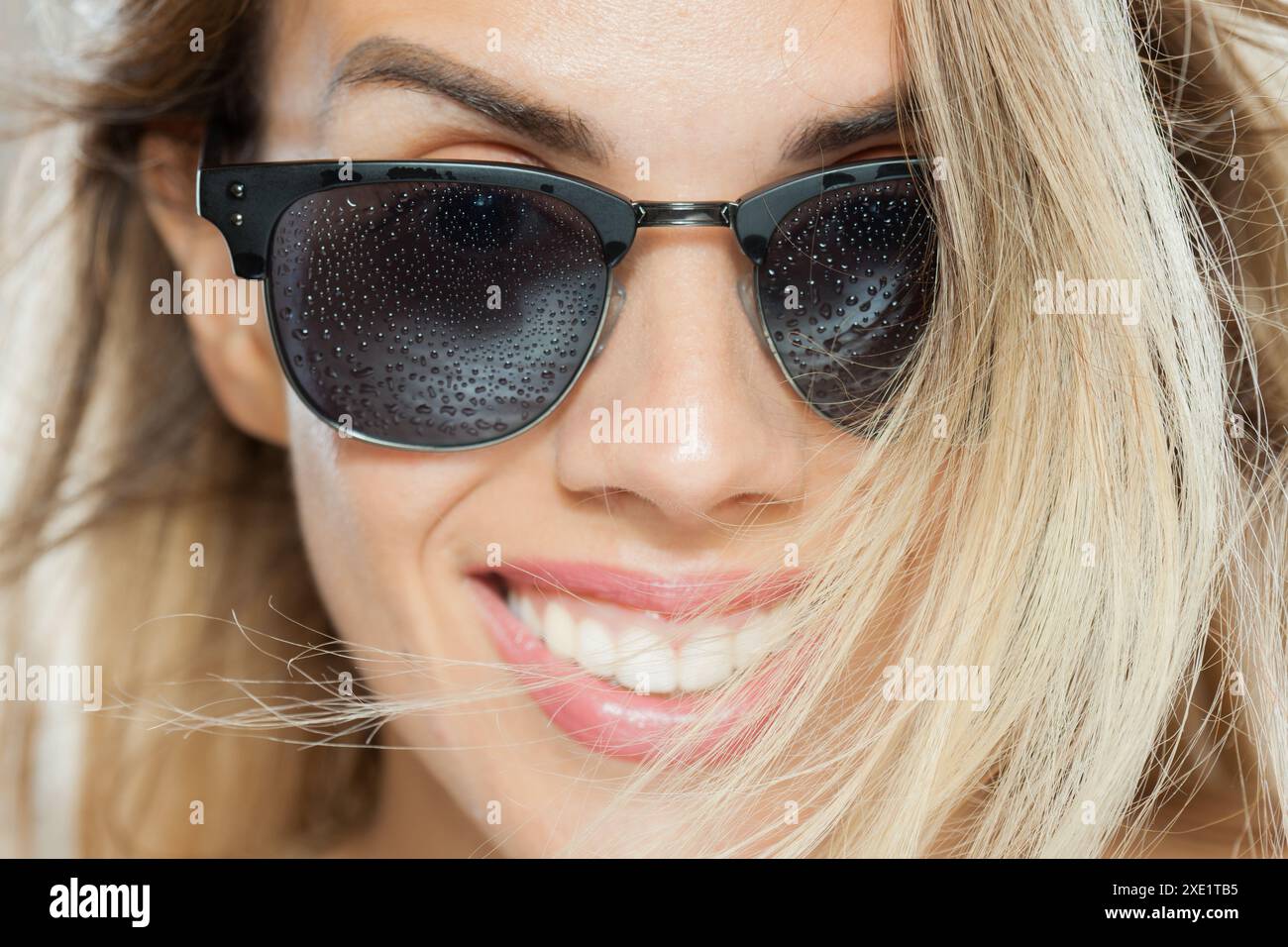 Un portrait vibrant capturant une femme joyeuse portant des lunettes de soleil tachetées de gouttes de rosée, mettant en valeur un moment ludique et embrassé par le soleil. Banque D'Images
