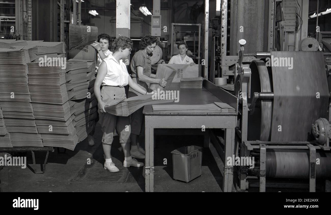 Années 1950, des travailleuses historiques triant des sacs d'épicerie en papier nouvellement produits, imprimés avec les mots 5-10 LB Cellos Potatoes, sur une table à côté d'une machine à imprimer, États-Unis. Banque D'Images