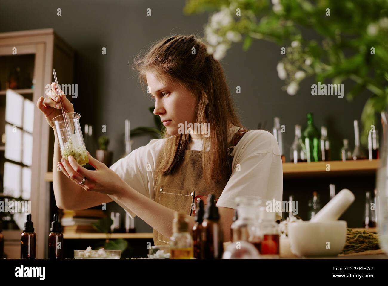 Jeune femme parfumeur assise au bureau de travail avec des articles d'artisanat de parfum et admirant le liquide doré avec des pétales de fleurs sauvages pendant qu'elle le mélange Banque D'Images