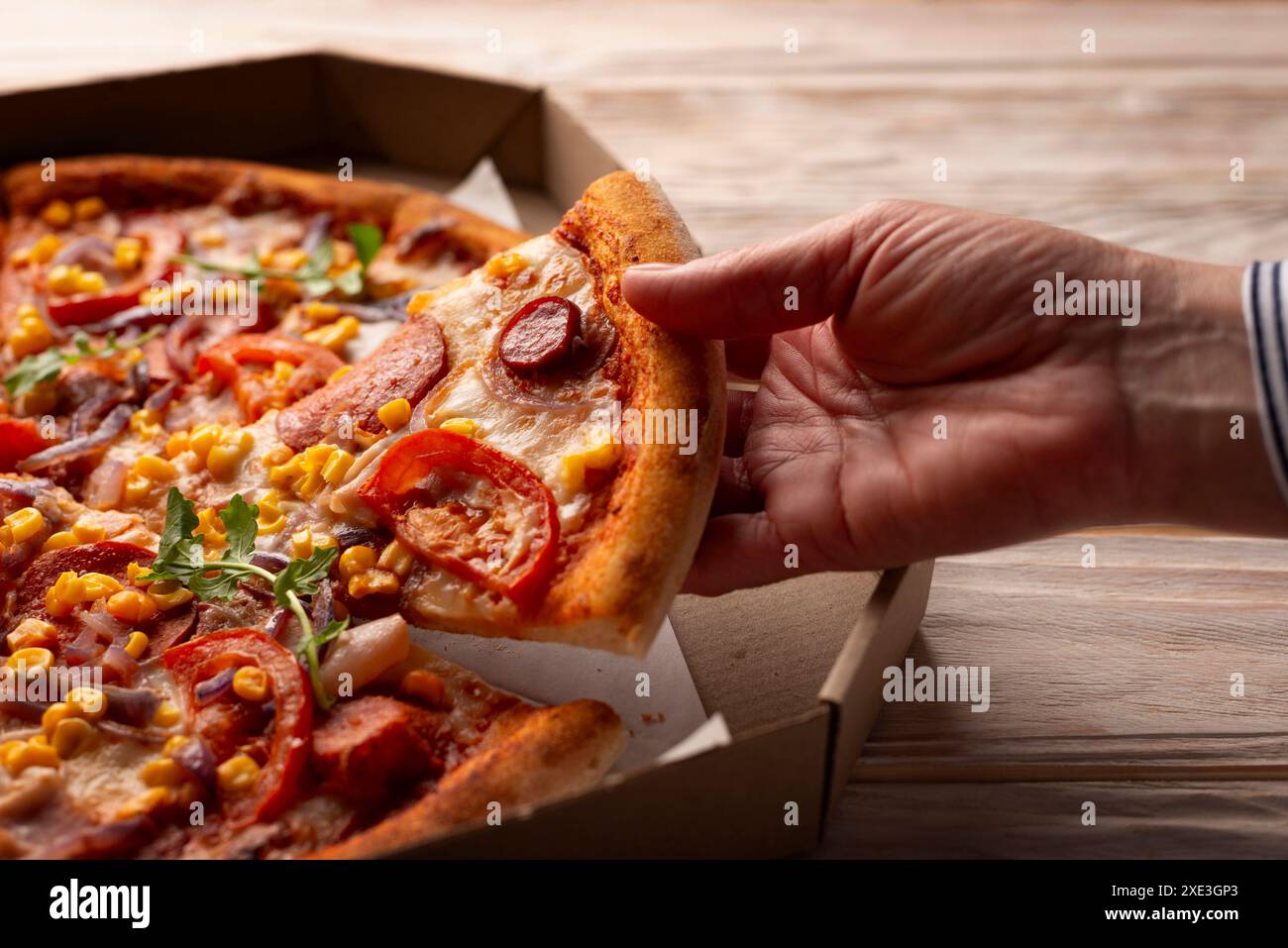 La main caucasienne humaine prend une tranche de grande pizza de la boîte de carton sur la table en bois blanc Banque D'Images