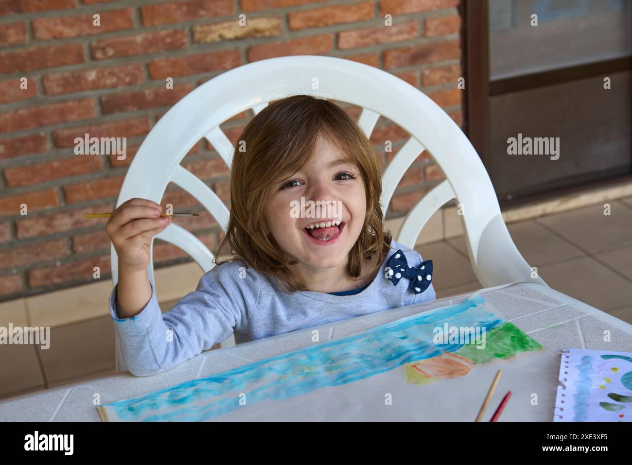 fille peignant un dessin d'aquarelle tout en tenant un pinceau dans sa main et souriant brillamment, avec un mur de briques en arrière-plan Banque D'Images