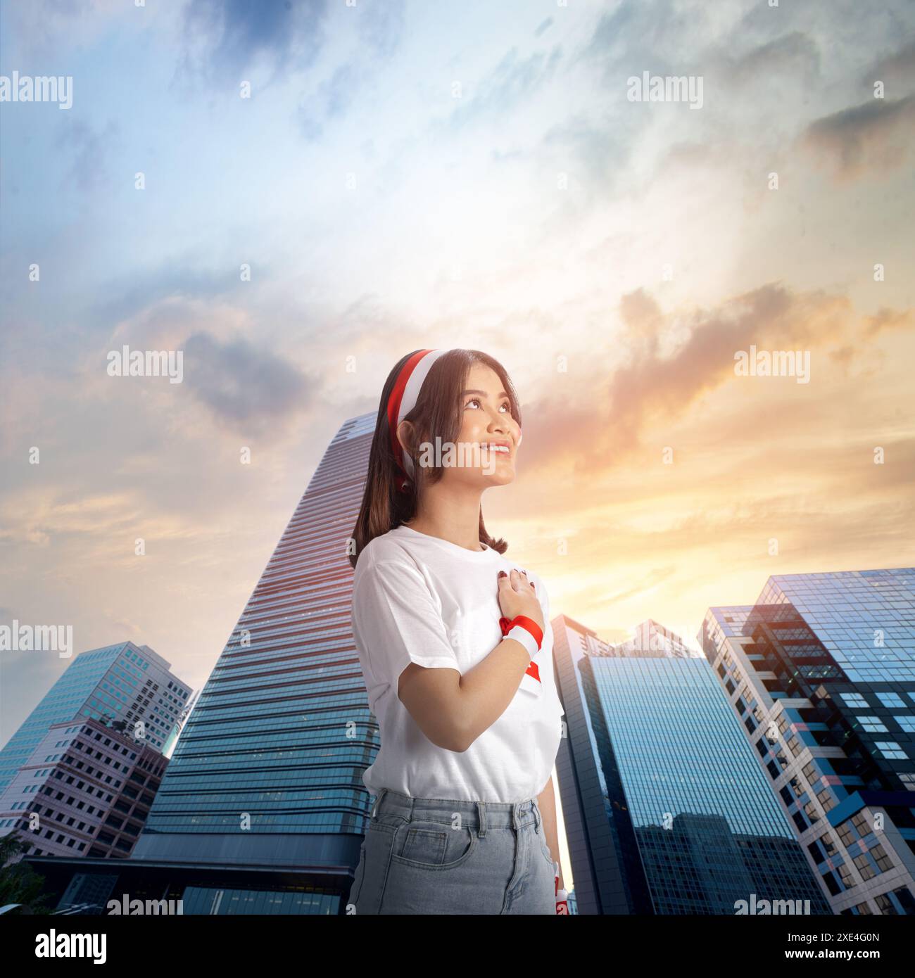 Jeune femme indonésienne pose sa main sur sa poitrine avec une expression fière, portant un bandeau de drapeau indonésien, dans une ville avec un beau soleil Banque D'Images