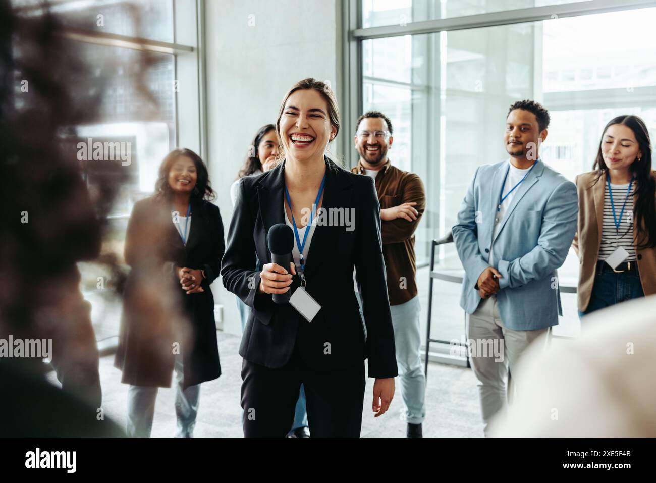 Femme d'affaires confiante avec microphone s'adressant à divers collègues dans un environnement de bureau moderne. Membres de l'équipe souriants et motivants pendant le pro Banque D'Images