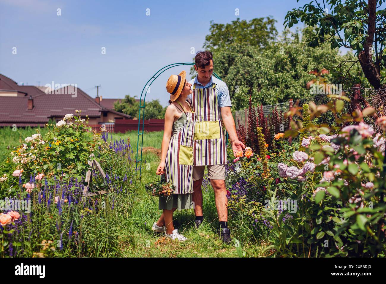 Heureux jeune couple appréciant les fleurs de roses marchant dans le jardin d'été. Jardiniers vérifiant les plantes portant des tabliers après le travail Banque D'Images