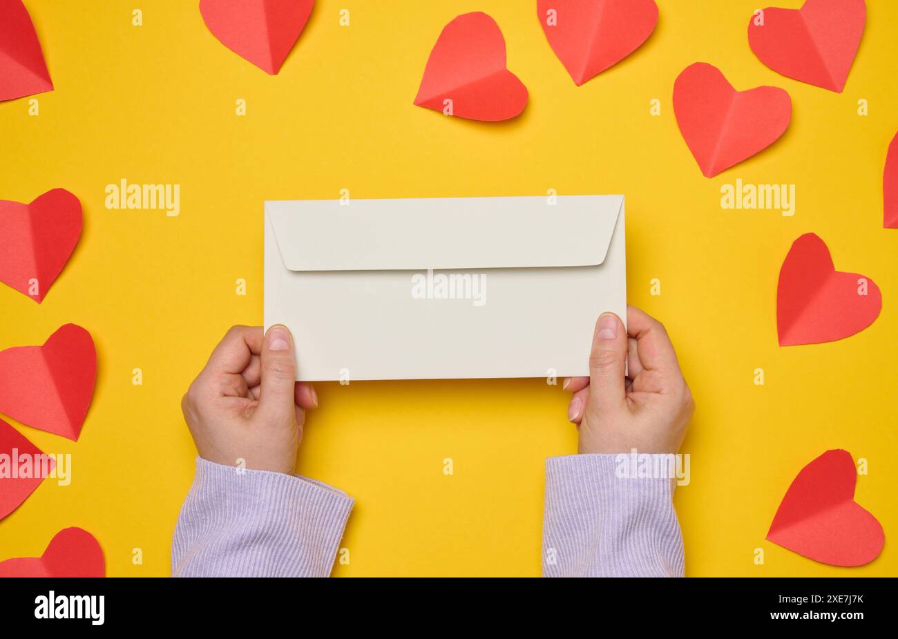 Les mains féminines tiennent une enveloppe rectangulaire blanche sur un fond jaune, des coeurs rouges en papier. Banque D'Images