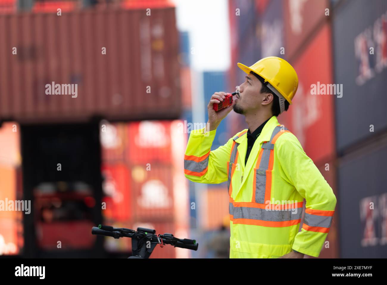 Portrait d'un ouvrier d'entrepôt parlant sur un talkie-walkie avec un collègue dans un entrepôt de conteneurs vide tout en conduisant un scoot Banque D'Images